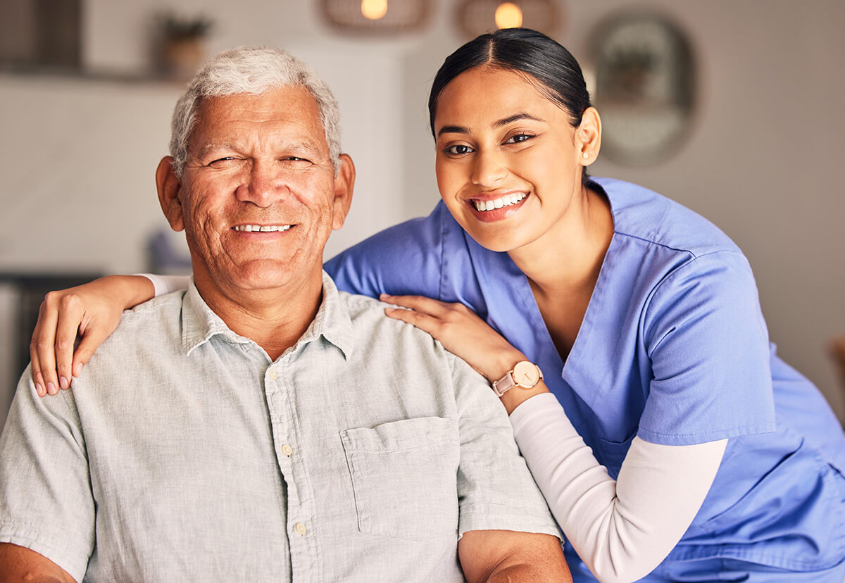 A professional caregiver in a light-filled home setting gently supports an older adult who is seated, offering companionship and assistance as they smile together.